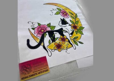 Hand-drawn black and white cat on gold crescent moon with pink and yellow floral embroidery pattern