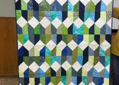 Handmade quilt in blue, green and white stacked diamond pattern held up at guild meeting