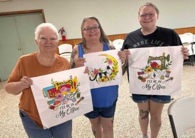 Three smiling quilt guild members holding hand-painted fabric panels with sewing machine and cat designs