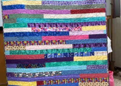 Smiling woman holding up a colorful handmade strip quilt at a guild meeting